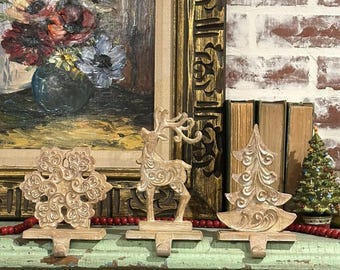 May include: Three beige Christmas stocking holders: a snowflake, a reindeer, and a Christmas tree. The holders are on a distressed green surface, with a red beaded garland. A framed painting and books are in the background.