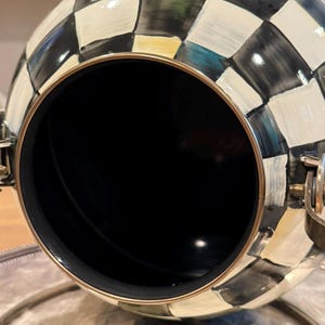 May include: A decorative, spherical container with a black interior and a checkered pattern in black, white, and gold. The container has a gold-colored rim and hinges, and is resting on a silver-colored surface.