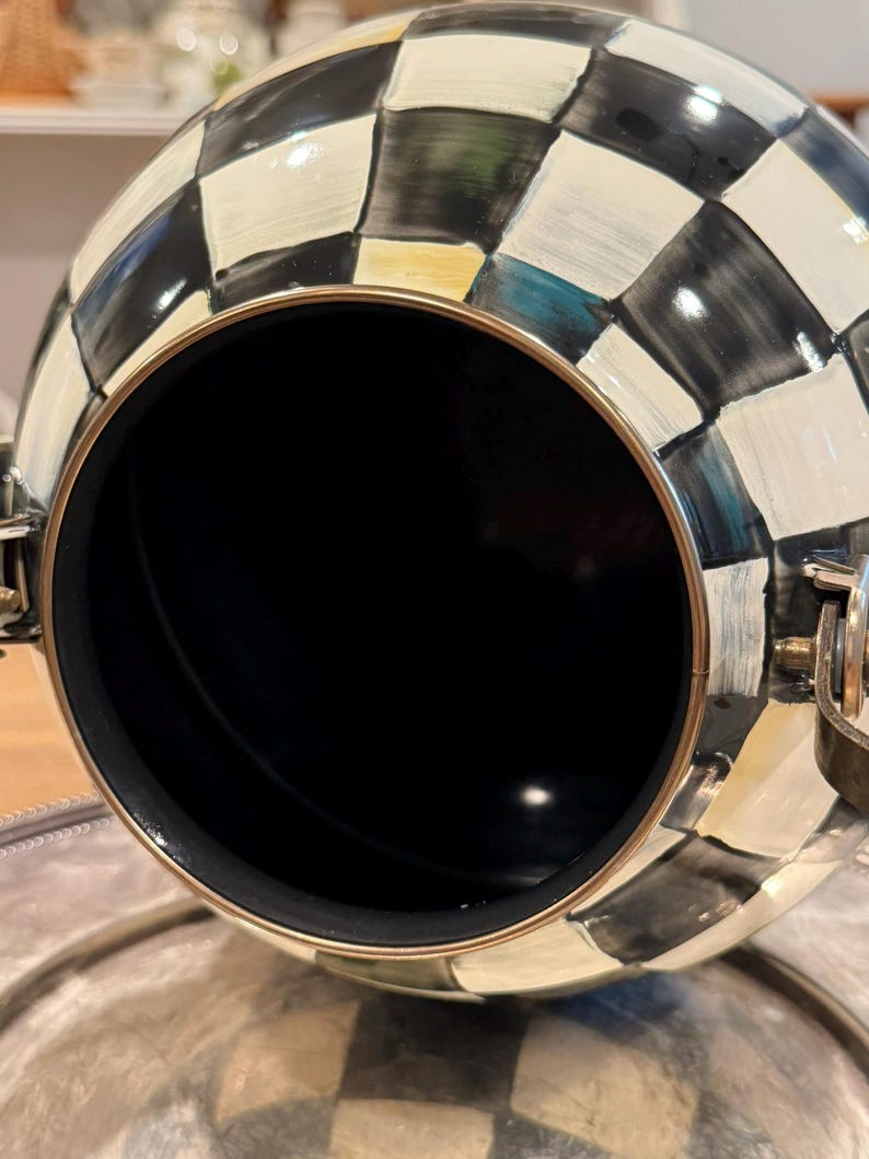 May include: A decorative, spherical container with a black interior and a checkered pattern in black, white, and gold. The container has a gold-colored rim and hinges, and is resting on a silver-colored surface.
