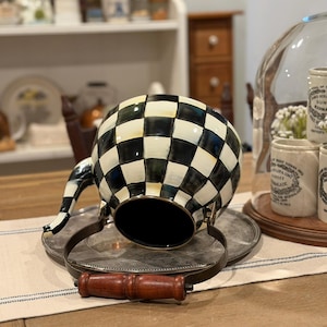 May include: A black and white checkered teapot with a silver metal handle and a wooden handle. The teapot is resting on a silver tray. The teapot has a black interior and a black and white striped spout.