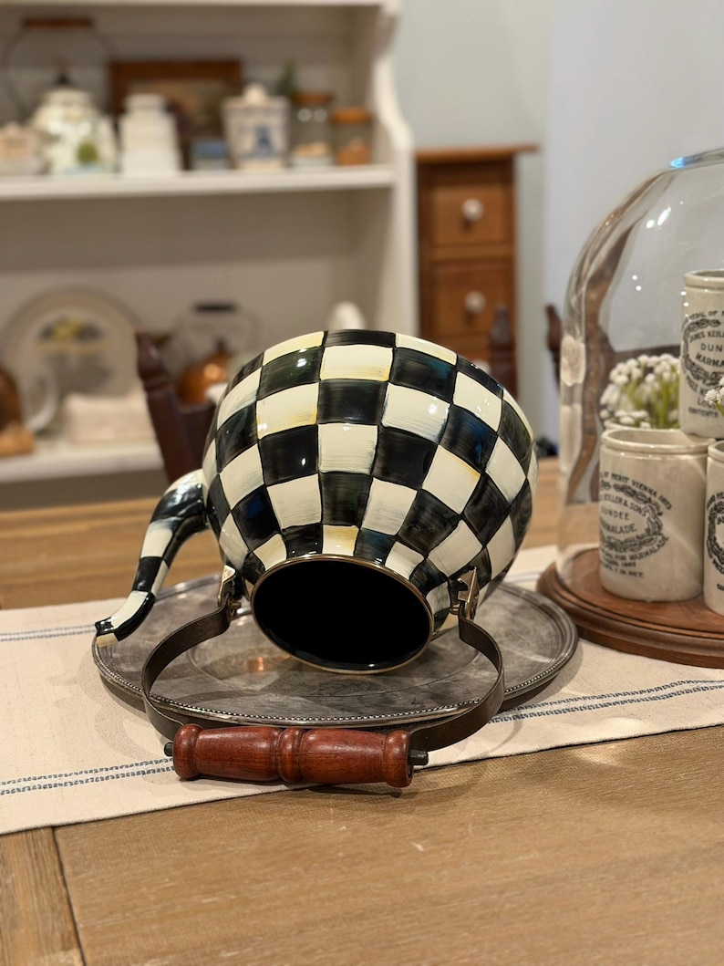 May include: A black and white checkered teapot with a silver metal handle and a wooden handle. The teapot is resting on a silver tray. The teapot has a black interior and a black and white striped spout.