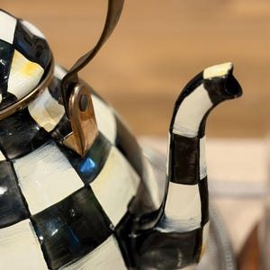 May include: A black and white checkered teapot with a curved spout and a handle. The teapot has a gold rim and a copper-colored bracket. The handle is black and curved. The teapot is on a silver tray.