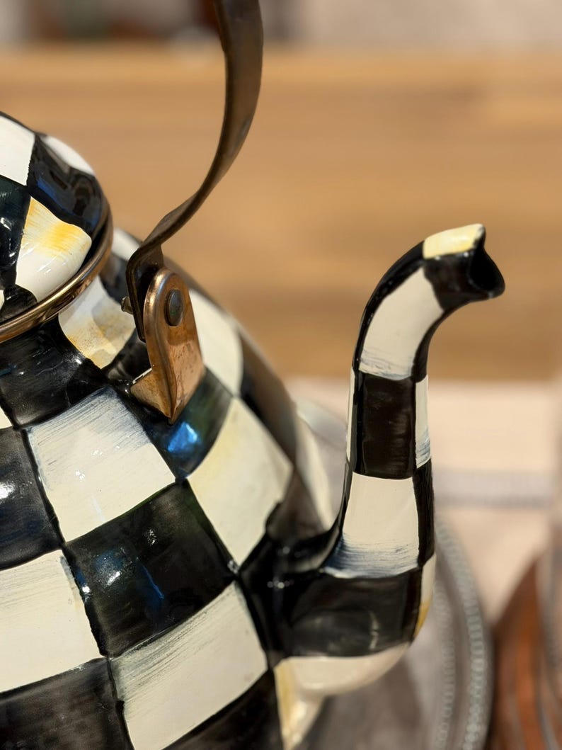 May include: A black and white checkered teapot with a curved spout and a handle. The teapot has a gold rim and a copper-colored bracket. The handle is black and curved. The teapot is on a silver tray.