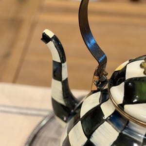 May include: A decorative black and white checkered teapot with a curved handle and spout. The teapot has a gold-colored rim and handle attachments. The spout is black and white striped.