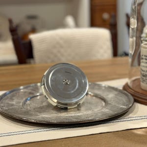 May include: A silver-colored, round serving platter with a beaded edge, paired with a matching lid. The lid is cylindrical with a silver top and a white and black band. The items rest on a striped placemat.