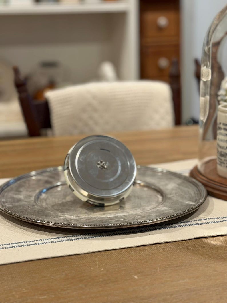 May include: A silver-colored, round serving platter with a beaded edge, paired with a matching lid. The lid is cylindrical with a silver top and a white and black band. The items rest on a striped placemat.