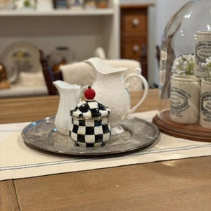 May include: A decorative arrangement featuring a silver tray with a small black and white checkered jar topped with a red apple, a white pitcher, and a smaller white creamer. A larger white pitcher with a dotted pattern is also present.
