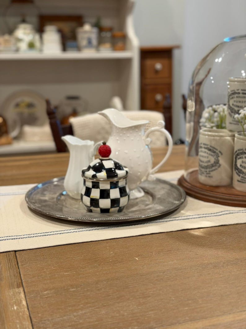 May include: A decorative arrangement featuring a silver tray with a small black and white checkered jar topped with a red apple, a white pitcher, and a smaller white creamer. A larger white pitcher with a dotted pattern is also present.