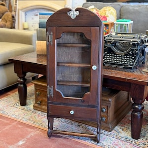 Op de afbeelding: Een donkerhouten kast met een glazen deur, een kleine lade en decoratieve hardware. De kast heeft een gebogen bovenkant en een handdoekstang aan de onderkant. Het is een vintage stuk, waarschijnlijk voor display of opslag.