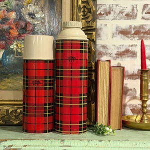 May include: Two vintage Thermos brand flasks with cream-colored lids. The flasks have a red and black plaid pattern with gold accents. The Thermos logo is visible on the front of each flask. One flask is taller than the other.