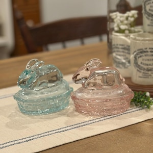 May include: Two glass bunny-shaped trinket boxes, one light blue and one pink, sit side-by-side on a striped table runner. The bunnies have detailed features and sit atop basket-like bases. Decorative items are in the background.
