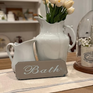 Może przedstawiać: Biały ceramiczny dzbanek z uchwytem trzyma bukiet kremowych tulipanów. Obok stoi mniejszy biały dzbanek. Prostokątny znak ze słowem "Bath" w skrypcie znajduje się przed dzbankami. Przedmioty znajdują się na paskowanym bieżniku.