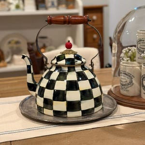 May include: A decorative black and white checkered teapot with a wooden handle and a red cherry-shaped finial. The teapot sits on a silver tray, suggesting a kitchen or dining room setting. The design is bold and eye-catching.