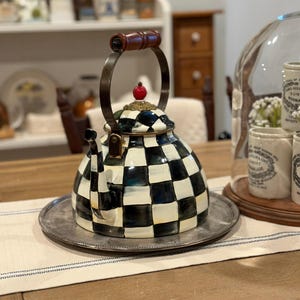 May include: A decorative black and white checkered teapot with a wooden handle and a red cherry-shaped finial. The teapot sits on a silver tray, suggesting a kitchen or dining setting.