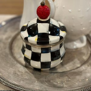 May include: A decorative sugar bowl with a black and white checkered pattern. The lid is topped with a red, textured finial. The bowl sits on a silver-colored plate, with white pitchers in the background.
