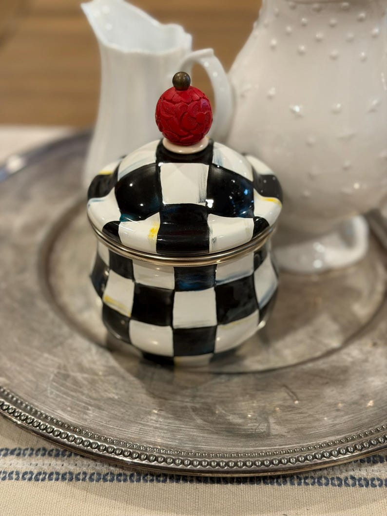 May include: A decorative sugar bowl with a black and white checkered pattern. The lid is topped with a red, textured finial. The bowl sits on a silver-colored plate, with white pitchers in the background.