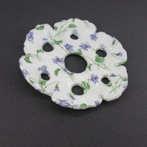 Puede incluir: Un objeto blanco, circular y texturizado con un borde festoneado y seis recortes circulares, que presenta un patrón repetitivo de pequeñas flores violetas con hojas y tallos verdes en toda la superficie de la pieza.