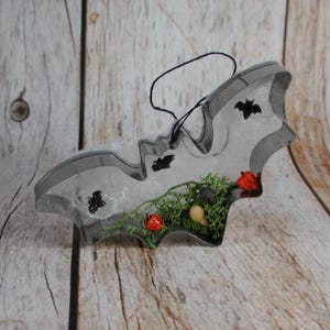 Puede incluir: Un cortador de galletas de metal con forma de murciélago relleno de resina transparente, que contiene musgo verde, pequeñas cuentas naranjas con forma de calabaza y diminutos detalles de murciélagos negros. Un hilo negro fino está unido en la parte superior.