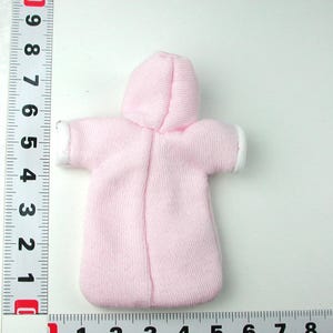 Puede incluir: Un conjunto de mu&ntilde;eca beb&eacute; de color rosa con capucha y ribete blanco alrededor de las mangas y la capucha. El conjunto est&aacute; hecho de un tejido suave y de punto.