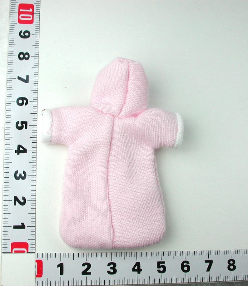 Puede incluir: Un conjunto de mu&ntilde;eca beb&eacute; de color rosa con capucha y ribete blanco alrededor de las mangas y la capucha. El conjunto est&aacute; hecho de un tejido suave y de punto.