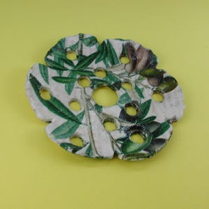 Puede incluir: Una jabonera circular de cerámica en color blanco roto con un estampado botánico de hojas de olivo verdes y aceitunas oscuras. La pieza presenta un borde festoneado irregular y múltiples orificios de drenaje circulares en su superficie.
