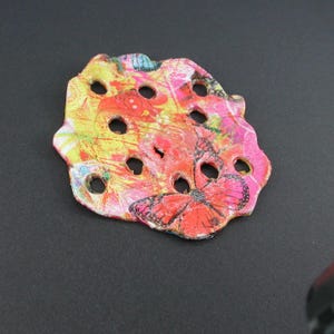 Puede incluir: Un objeto decorativo plano de forma irregular con una superficie vibrante y multicolor que presenta salpicaduras de amarillo, naranja y rosa. La pieza tiene varios recortes circulares y un gráfico de mariposa con bordes negros.