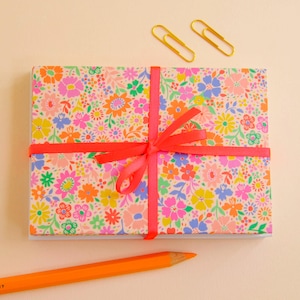 May include: A rectangular stack of paper featuring a vibrant floral pattern in shades of pink, orange, yellow, blue, and green. The stack is tied with a bright orange ribbon in a bow. Two gold paper clips and an orange pencil are nearby.