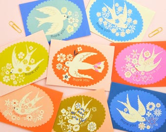 Puede incluir: Una colección de tarjetas de agradecimiento coloridas. Cada tarjeta presenta un diseño de pájaro blanco dentro de un marco ovalado, rodeado de flores. Las tarjetas son de varios colores, incluyendo azul, naranja, rosa y verde, con la frase "gracias" impresa en cada una.