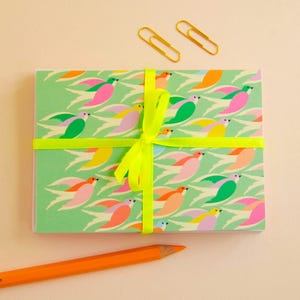 Peut inclure: Une pile de cartes rectangulaires présentant un motif répétitif d'oiseaux stylisés dans des tons de rose, orange, jaune et vert sur un fond vert clair, nouées avec un ruban jaune vif, accompagnées de deux trombones dorés et d'un crayon orange.