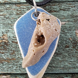 May include: A blue and white ceramic pendant with a beige, porous rock attached. The pendant is on a black braided cord.