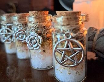 May include: Four small glass jars with cork tops filled with white crystals and dried herbs. Each jar has a silver charm hanging from the cork, including a pentagram and a flower.