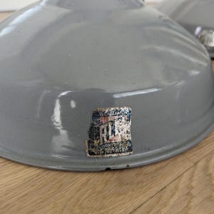 May include: Two gray, industrial-style pendant lamps with a glossy finish. Each lamp has a wide, rounded shade. A faded label is affixed to the front of one lamp. The lamps are suspended from chains and are likely for overhead lighting.