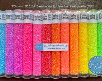 May include: Twelve tubes of 11/0 size DELICA luminous beads in various colors, including blue, pink, orange, yellow, and green. The tubes are labeled "Little Spirit Beads.com". The text "(12) Colors. DELICA Luminous size 11/0 beads in 3 IN Standard/TBS" is visible at the top of the image. The text "Little Spirit Beads LLC Wisconsin Shop Est. 2013" is visible in the center of the image. The text "Little Spirit Beads" is visible in a blue circle at the bottom of the image.