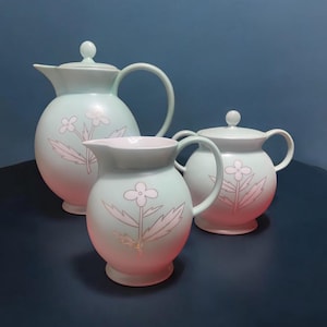 May include: A set of three pastel green ceramic teapots with white floral designs. The teapots have a gradient design with a pink base.