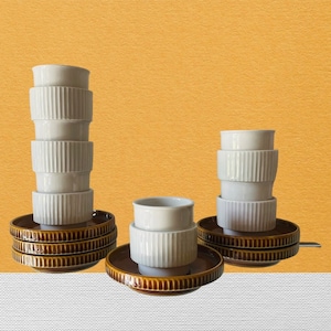 Può includere: Un set di sei tazze in ceramica bianca con motivi a coste, impilate in tre coppie. Ogni coppia è su un piattino marrone con un bordo rialzato. I piattini sono impilati uno sopra l'altro.