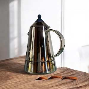 Può includere: Una macchina per caffè espresso da piano cottura in argento e blu con una manopola blu sul coperchio. La macchina è appoggiata su una superficie in legno.