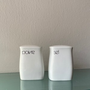 Può includere: Due saliere e pepiere in ceramica bianca con scritta nera. La saliera a sinistra dice "poivre" e la pepiera a destra dice "sel".
