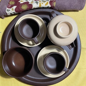 Può includere: Un vassoio in ceramica marrone con quattro tazze e piattini in ceramica beige. Le tazze hanno un interno marrone e i piattini hanno un bordo marrone.