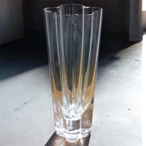 May include: A tall, clear glass vase with a unique, fluted design. The vase has a wide, scalloped top and a base with three rounded feet. The transparent glass allows light to pass through, creating shadows. A decorative piece for floral arrangements.