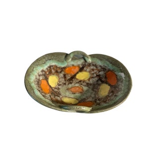 Può includere: Una ciotola in ceramica verde con un manico. La ciotola ha una glassa macchiettata ed è decorata con macchie arancioni e gialle.