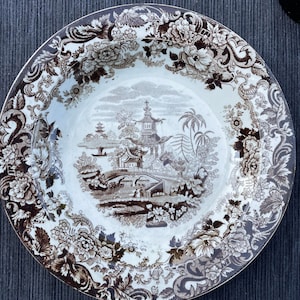 Può includere: Una ciotola in ceramica bianca con un motivo floreale marrone e bianco attorno al bordo. Il centro della ciotola presenta un'illustrazione marrone e bianca di una pagoda e un ponte in un paesaggio.
