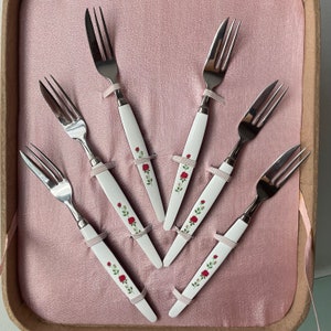May include: Set of six silver cake forks with white handles decorated with red roses. The forks are arranged in a fan shape in a pink box.