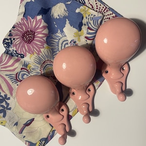 Può includere: Tre attaccapanni da parete rosa pastello con una parte superiore rotonda e un design decorativo. Gli attaccapanni sono montati su una superficie bianca, con un tessuto a fantasia floreale sullo sfondo. Gli attaccapanni sono di colore rosa tenue.