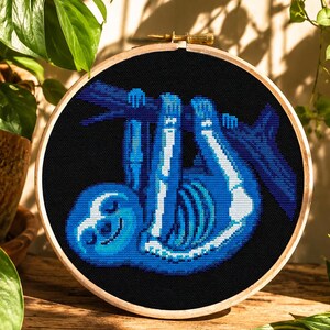 May include: A cross-stitch embroidery hoop with a blue sloth skeleton design on a black background. The sloth is hanging from a branch, with a wooden hoop and green plants in the background.