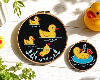 May include: Two cross-stitch hoops with yellow duck designs on black fabric, alongside two rubber duck toys. One hoop shows ducks swimming, the other shows a duck relaxing with a drink. The hoops are framed in wooden embroidery hoops.