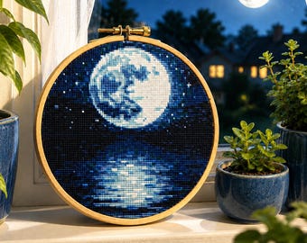 Könnte beinhalten: Ein runder Stickrahmen aus Holz mit einem Kreuzstichmuster, das einen Vollmond in einem dunkelblauen Nachthimmel zeigt, wobei sich der Mond auf einer dunklen Wasseroberfläche spiegelt, vor einem Hintergrund mit einem echten Nachthimmel.