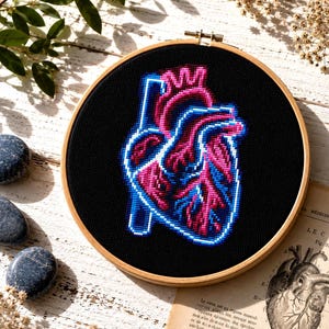 May include: A cross-stitch embroidery of a human heart in pink and blue against a black background, set in a wooden hoop. The design is on a white wooden surface with decorative stones and dried flowers.