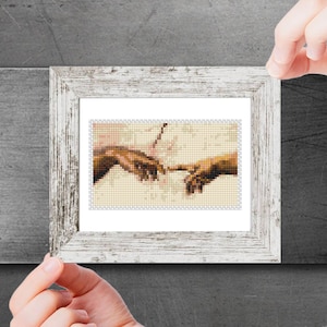 Puede incluir: Una obra de arte de punto de cruz enmarcada que representa un detalle de La Creación de Adán de Miguel Ángel, que muestra dos manos que se acercan.