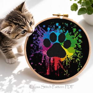 Op de afbeelding: Een kruissteekpatroon in een houten hoepel toont een zwarte pootafdruk met een kleurrijk regenboogspatterontwerp op een zwarte achtergrond. De tekst "Cross Stitch Pattern PDF" staat onderaan. Een katje kijkt naar de kruissteek.