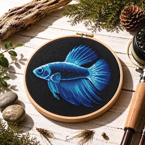 Puede incluir: Un bastidor de bordado de punto de cruz con un pez betta azul vibrante sobre un fondo negro. El pez está detallado con diferentes tonos de azul, en un bastidor de madera. La imagen está rodeada de aparejos de pesca y elementos naturales.